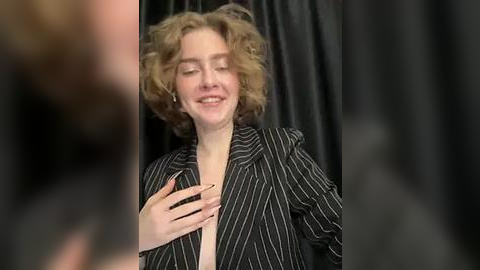 Media: Video of a smiling, fair-skinned woman with curly blonde hair, wearing a black pinstriped blouse, standing against black curtains. She is touching her chest, her nails painted black.