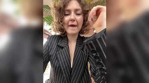 Media: A candid video captures a woman with curly hair in a pinstriped black blazer, adjusting her hair, against a blurred background of greenery and a building.