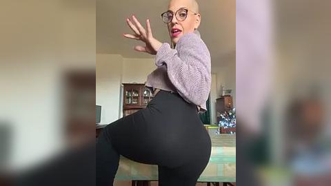 Media: A video of a bald, middle-aged woman with glasses, wearing a light purple sweater and black leggings, mid-dance in a dimly lit living room.