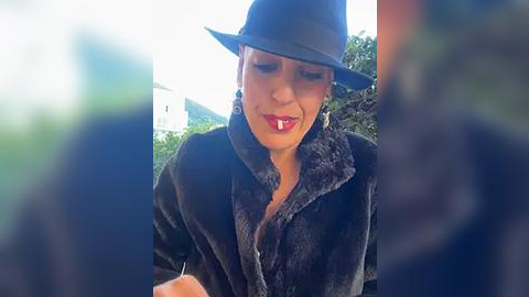 Media: A video shows a woman in a dark fur coat and fedora, smiling with her mouth open, holding a small object. The background features blurred greenery and a white building.