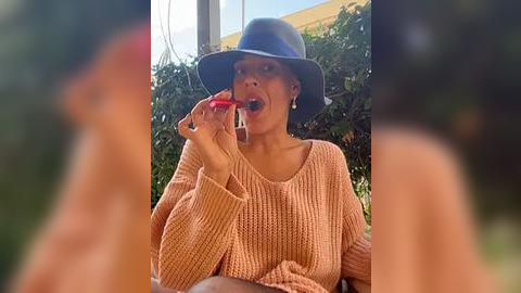 Media: A video of a woman with a medium-dark complexion wearing a light peach knitted sweater and a dark blue hat, smiling and holding a red object in her mouth, set outdoors with a blurred background.