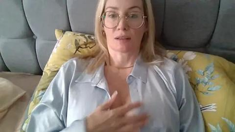 Media: Video of a middle-aged Caucasian woman with blonde hair and glasses, wearing a light blue blouse, lying on a grey couch with yellow floral pillows.