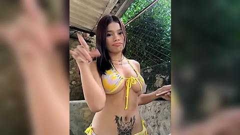 Media: Video of a young Asian woman with long black hair, wearing a yellow bikini, making a peace sign, in a rustic outdoor setting with a chain-link fence and stone wall.