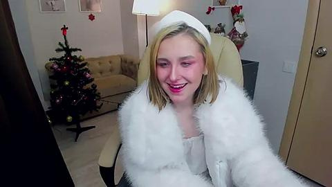 Media: A video of a smiling, blonde woman in a white fur coat and hat, sitting on a beige chair in a cozy, festive living room with a Christmas tree and decorated shelves.