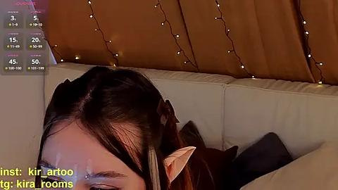 Media: A video of a person with long, dark hair and pointy ears, lying on a beige couch with a brown curtain backdrop and fairy lights. Text at the bottom reads, \"First: kirara to kirara rooms.\