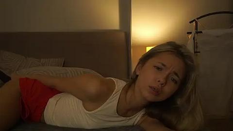 Media: Video of a young woman with light skin and blonde hair, wearing a white tank top and red shorts, lying on a bed, looking worried.