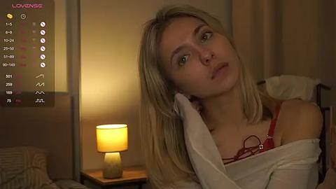 Media: Video of a blonde woman with fair skin, wearing a red bra, leaning on a bed, illuminated by a bedside lamp, in a dimly lit bedroom.