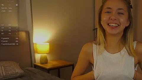 Media: Video of a smiling young woman with light skin and blonde hair, wearing a sleeveless white top, standing in a dimly-lit, minimalist bedroom with a wooden nightstand and lamp.