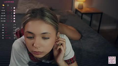 Media: Video of a young woman with light skin and brown hair, lying on her stomach on a bed with a grey blanket, wearing a white shirt. Background includes a nightstand with a lamp, a mirror, and a window.