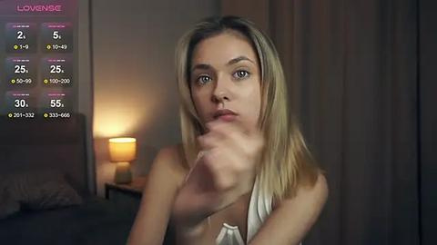 Media: Video of a young woman with straight blonde hair, fair skin, and a slim build, wearing a white halter top, looking pensively at her phone screen, which shows a \"Lomake\" app with various options. Background includes a dimly lit room with a bedside lamp and a closed wardrobe.