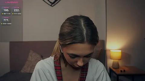 Media: A video of a young woman with straight, shoulder-length brown hair, wearing a red plaid shirt, sitting on a bed with a beige pillow. The background includes a wooden nightstand with a lamp and a wall clock.