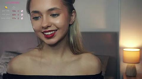 Media: Video of a young woman with light skin, blonde hair, and red lipstick, smiling in a dimly-lit bedroom with a beige headboard and a lamp.