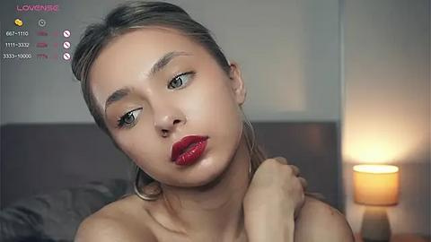 Media: Video of a light-skinned woman with long, straight blonde hair and striking red lipstick, wearing a strapless top. Background includes a lamp and blurred couch.