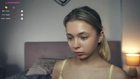 Media: Video of a young, light-skinned woman with blonde hair, wearing a yellow tank top, sitting on a bed in a dimly lit room.