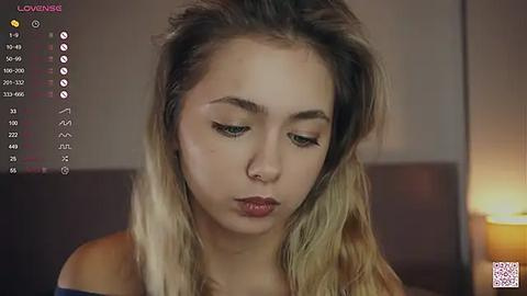 Media: Video of a young woman with long, wavy, blonde hair, wearing an off-shoulder top, looking down thoughtfully. Background shows a dimly lit room with a lamp on the right.