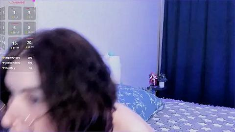 Media: A blurry video of a woman with curly dark hair, lying on a bed with a blue and white star-patterned blanket. A small, stuffed toy is on the right.