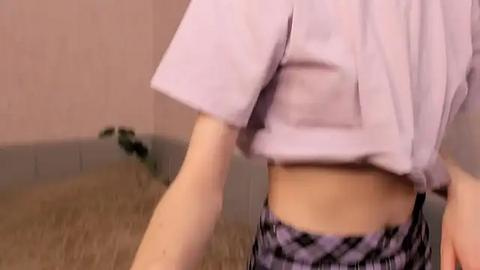 Media: Video of a person in a light purple cropped top, with a black and white patterned skirt, standing in a room with beige carpet and light pink walls.