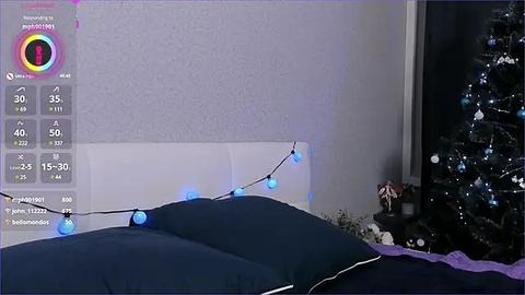 Media: Video of a cozy bedroom at night, featuring a dark bed, a glowing string of blue lights, a snow-covered Christmas tree, and a white wall.