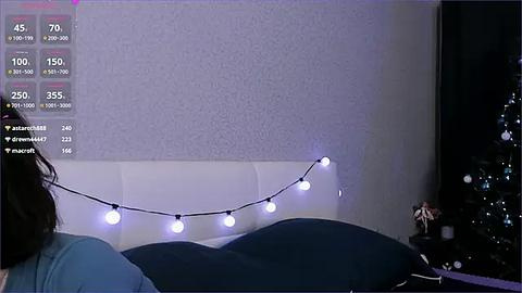 Media: Video of a dimly lit bedroom with a white headboard, dark bedding, and a string of white fairy lights. A woman with dark hair, wearing a blue shirt, lies on the bed. The background features a textured, light purple wall and a partially visible Christmas tree.