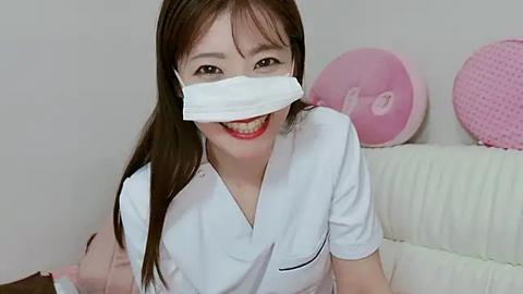 Media: Video of a smiling Asian woman in a white nurse uniform with a white face mask, seated on a white couch. Background features a pink cushion and a white wall.