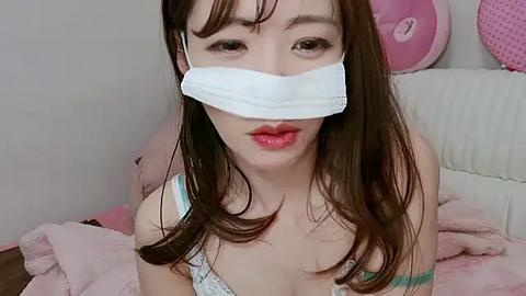 Media: Video of a young Asian woman with long brown hair, wearing a white face mask, pink lipstick, and a light blue bra, sitting on a pink blanket in a softly lit room.