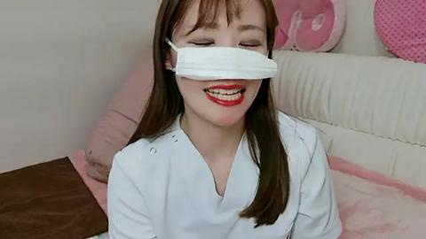 Media: Video of a smiling Asian woman with straight brown hair, wearing a white surgical mask and a white scrub top, sitting on a pink bed with a white pillow.