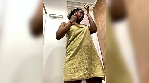 Media: Video of a dark-skinned woman with braids, wrapped in a yellow towel, taking a selfie in a small, dimly lit bathroom with white walls and a wooden door.