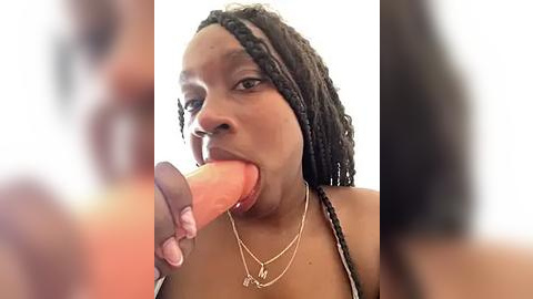 Media: Video of a dark-skinned Black woman with braided hair, wearing a necklace, sucking a large, erect penis. She has a neutral expression, and the background is out of focus.