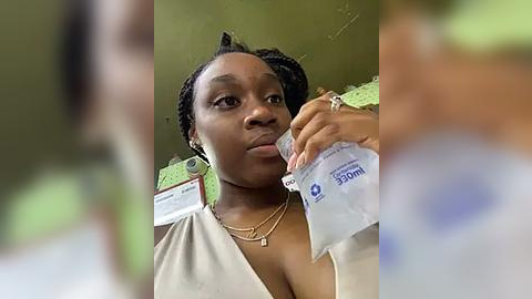 Media: Video of a young Black woman with braided hair, wearing a beige dress, drinking from a white water bottle, with a blurred background showing a green wall and green paper.