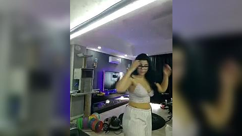 Media: A video of a young woman with long black hair, wearing a white crop top and high-waisted pants, taking a selfie in a dimly lit, cluttered living room with a flat-screen TV, a shelf, and a cooler on the floor.