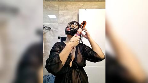 Media: Video of a woman in a black latex mask and robe, applying makeup in a modern bathroom with grey tiles and a white door.