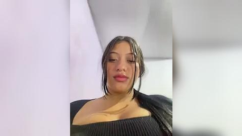 Media: Video of a Latina woman with medium skin tone, dark hair, and full lips, wearing a black, ribbed sweater, standing in a modern, white-walled room.