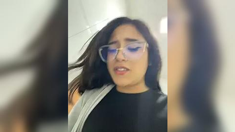 Media: A video of a young woman with dark hair, wearing large glasses, a black top, and a gray backpack, appearing startled, in a blurred, indoor setting.