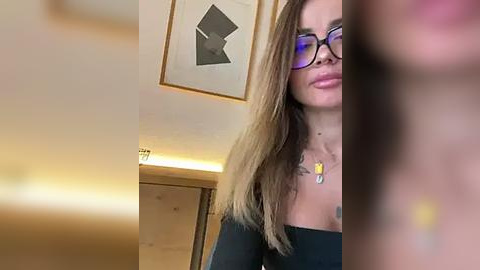 Media: Video of a woman with long, straight, light brown hair, wearing black-rimmed glasses and a black off-shoulder top, standing indoors near a framed abstract art piece on a light-colored wall.