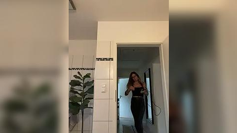 Media: Video of a woman with long brown hair taking a mirror selfie in a modern, light-toned bathroom. She wears a black tank top and pants, standing in front of a white-tiled wall with a small plant in the background.