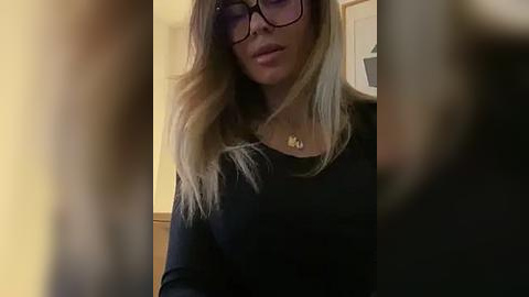 Media: Video of a woman with long, wavy blonde hair, wearing black-framed glasses and a black top, with a blurred background featuring a beige wall and a white door.