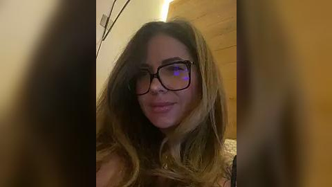 Media: Video of a fair-skinned woman with long, wavy brown hair, wearing black-rimmed glasses and a subtle smile, in a dimly lit room with wooden walls and a bed in the background.