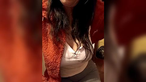 Media: A video of a woman with long, dark hair, wearing a white camisole and grey pants, sitting in a cozy room with red and brown hues.