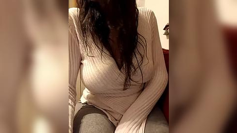 Media: Video of a woman with long, wet hair, wearing a snug, ribbed, beige sweater, sitting on a carpeted floor. The background is blurred, focusing on her torso and upper legs.