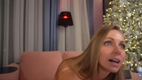 Media: Video of a blonde woman with light skin, sitting on a pink couch, gazing to the right. Background includes a lit Christmas tree, draped curtains, and a lamp with a red shade.