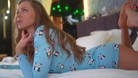 Media: Video of a young Caucasian woman with long brown hair, lying on her stomach in a blue onesie with panda patterns, on a bed with white linens, in a cozy, dimly lit room with green fairy lights.