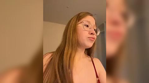 Media: Video of a young Caucasian woman with long, straight, light brown hair and glasses, wearing a red top, standing indoors with beige walls.