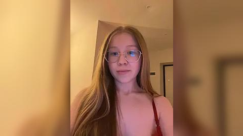 Media: Video of a young, fair-skinned woman with long, straight blonde hair, wearing glasses and a red top, standing indoors against a beige wall with a door and light fixtures in the background.