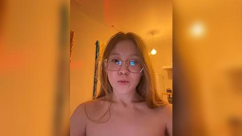 Media: Video of a young, topless woman with light skin, straight blonde hair, and glasses, in a warmly lit room with yellow walls and hanging art.