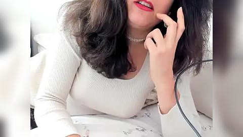 Media: Video of a woman with medium skin tone, dark curly hair, wearing a white ribbed long-sleeve top and a choker necklace, holding a smartphone to her mouth, on a bed with white floral-patterned sheets.