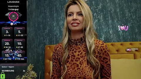 Media: Video of a smiling, blonde woman with long, wavy hair, wearing a leopard-print top, seated in a mustard-yellow upholstered chair against a textured teal wall. A game interface with a \"Love Meter\" is displayed on the left.