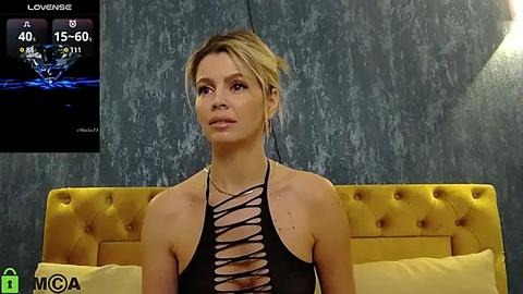 Media: Video of a blonde woman with fair skin wearing a revealing black dress with cutouts, seated on a mustard-yellow tufted headboard in a dimly lit room with textured gray walls.