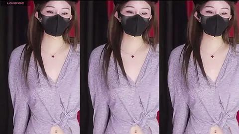 Media: A video of a young woman with long brown hair, wearing a black face mask and a grey, tie-dye V-neck top, standing against a black and red curtain background.