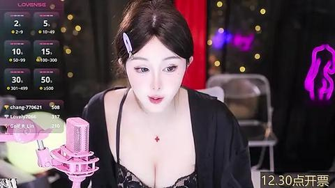 Media: Video of a young East Asian woman with fair skin, black hair in a bun, and a black lace bra, holding a pink microphone, in a dimly lit room with blurred background.