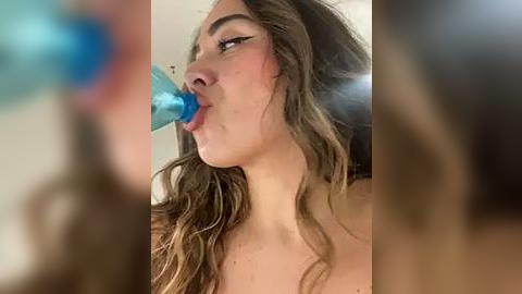 Media: Video of a young woman with wavy brown hair, fair skin, and blue lipstick, drinking from a blue straw. Background is blurred and indistinct.